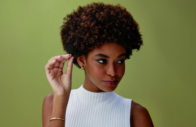 Foto de una mujer con cabello crespo corto en tonos castaños. El fondo es verde, y lleva una blusa blanca sin mangas mientras sostiene un mechón de su cabello con la mano izquierda.