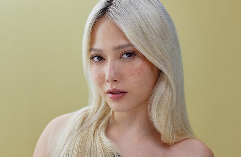 Foto de una mujer con cabello liso, largo y rubio, peinado hacia un lado. El fondo es amarillo claro, y lleva una blusa negra de un solo hombro.