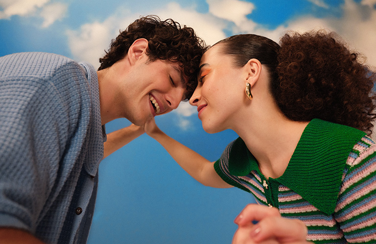 Toma plano medio de una pareja de hombre y mujer tomados de la mano y juntando sus frentes. Él tiene el cabello corto y ella rizado recogido.
