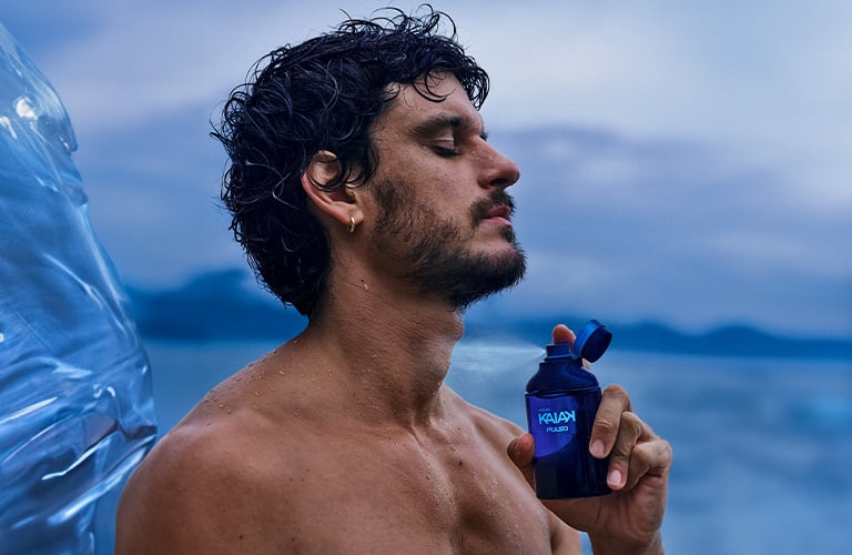 Un hombre sin camisa, con los ojos cerrados, rocía un perfume azul de la marca Kaiak. Fondo azul claro.