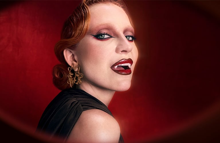 Foto con fondo rojo oscuro. Mujer con cabello recogido, accesorios dorados en la oreja derecha, maquillaje estilo vampiro con labios rojos intensos y ojos oscuros.