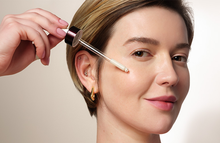 Close up modelo sosteniendo un gotero con sérum sobre su mejilla.