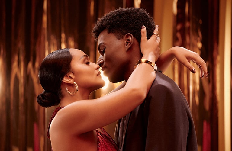 Close up de una pareja de hombre y mujer, abrazados y casi besándose. Ella tiene el cabello recogido y él un pequeño afro. Ambos vestidos elegantes.