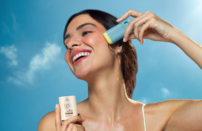 Foto de persona al aire libre con cielo azul de fondo; sostiene un protector solar en barra Natura Solar  en una mano y otro envase cerca de la sien, con enfoque en cuello y hombros.