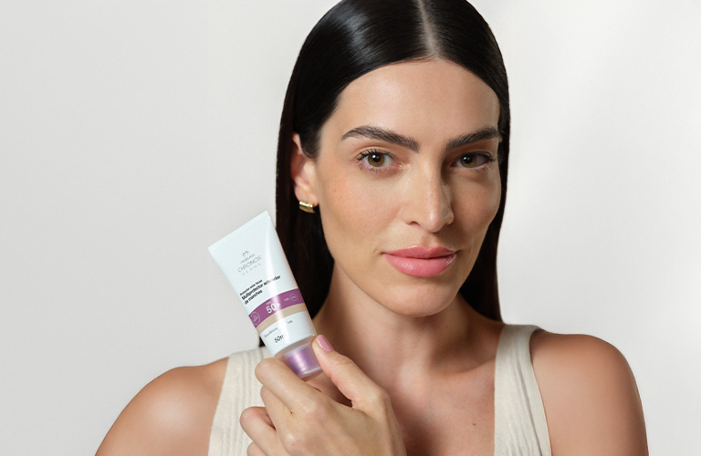 Foto con fondo claro. Persona sostiene un tubo de protector facial Chronos Derma FPS 50 frente al pecho; se ven mano, aro en la oreja y parte de un top claro.
