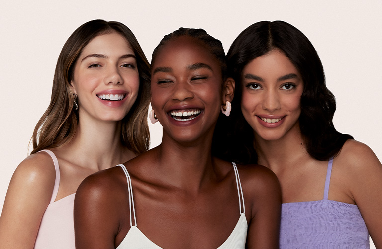 Foto de tres mujeres con diferentes tipos de cabello. El fondo es beige claro, y llevan ropa en tonos pastel: rosa, blanco y lila.