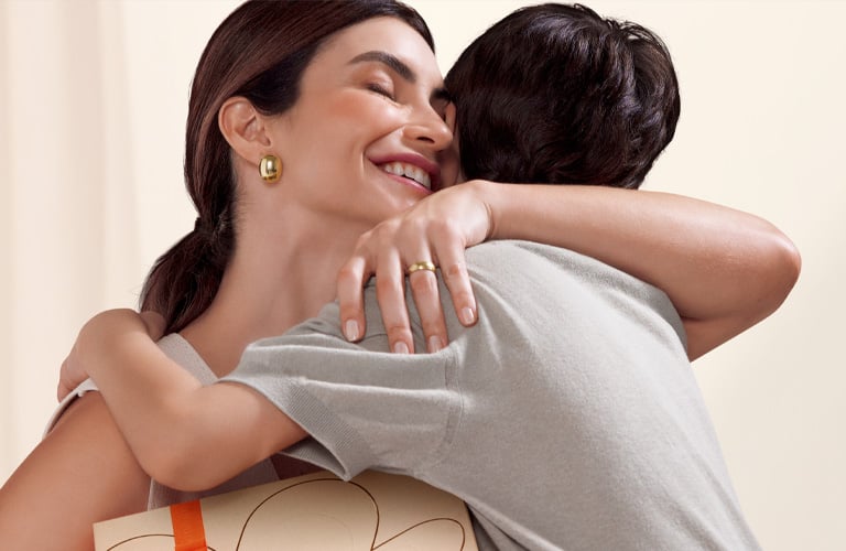 Fondo claro. Una mujer sonriente, con los ojos cerrados, abraza a un niño. La mujer lleva pendientes y un anillo. Se ve un regalo con cinta naranja.