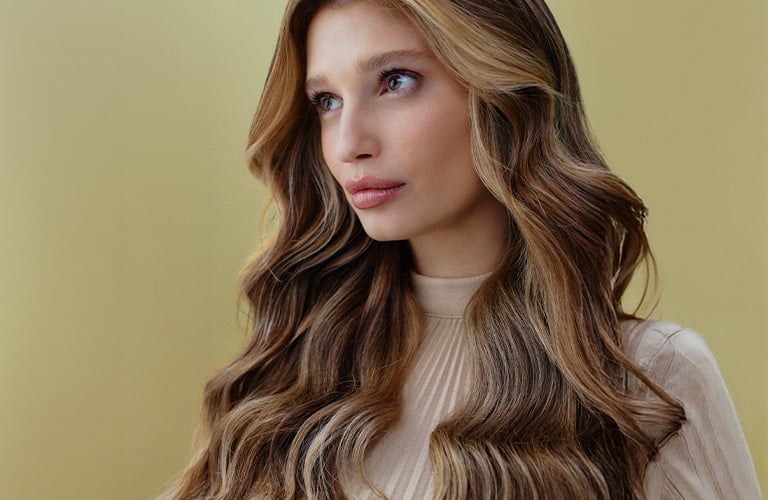 Foto de una mujer con cabello ondulado, largo y con mechas en tonos castaños y rubios. El fondo es amarillo claro, y lleva una blusa beige con textura delicada.
