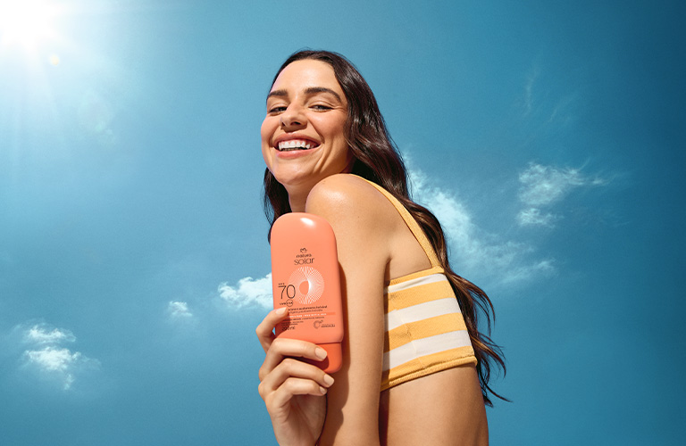 Foto con fondo de cielo azul con sol y nubes. Persona con top a rayas sostiene un protector solar FPS 70 color coral junto al hombro. Enfoque en el producto y la luz.