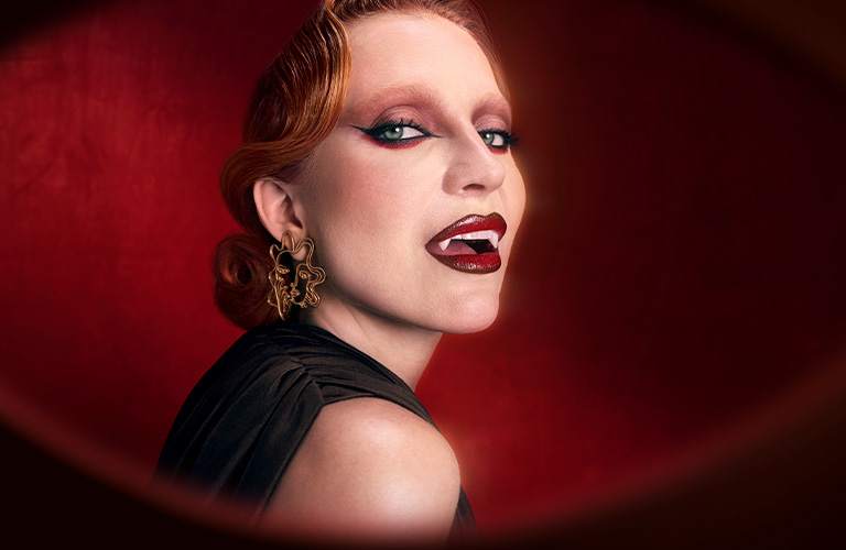 Foto con fondo rojo oscuro. Mujer con cabello recogido, accesorios dorados en la oreja derecha, maquillaje estilo vampiro con labios rojos intensos y ojos oscuros.
